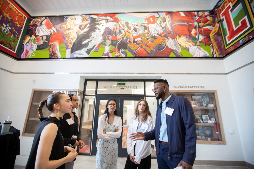 Stewy Robertson '11 H'18 speaks ot his art students in the Battle-Fitzgerald Atrium as their colorful, multimedia mural wraps around the three walls around them, near the ceiling.