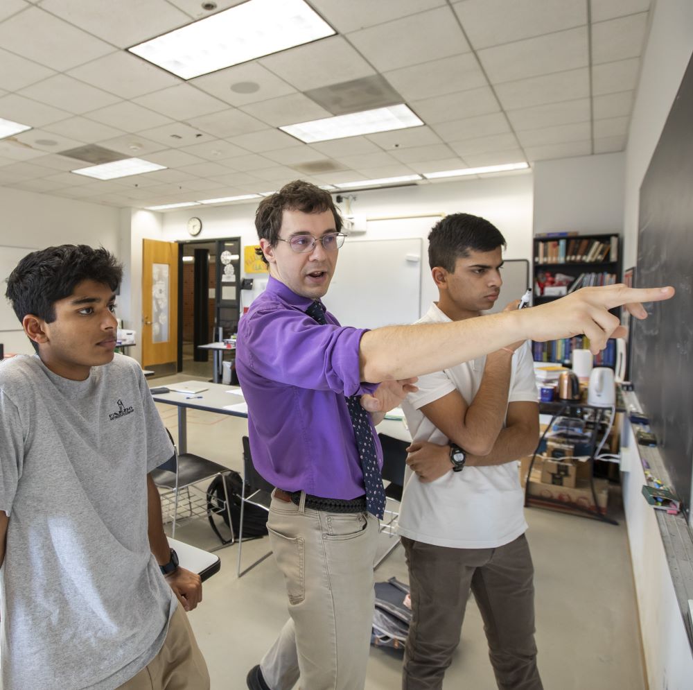 Math teacher Anton Fleissner, wearing a purple dress short with the sleeves rolled up, points to an equation on the chalkboard while two students look on.