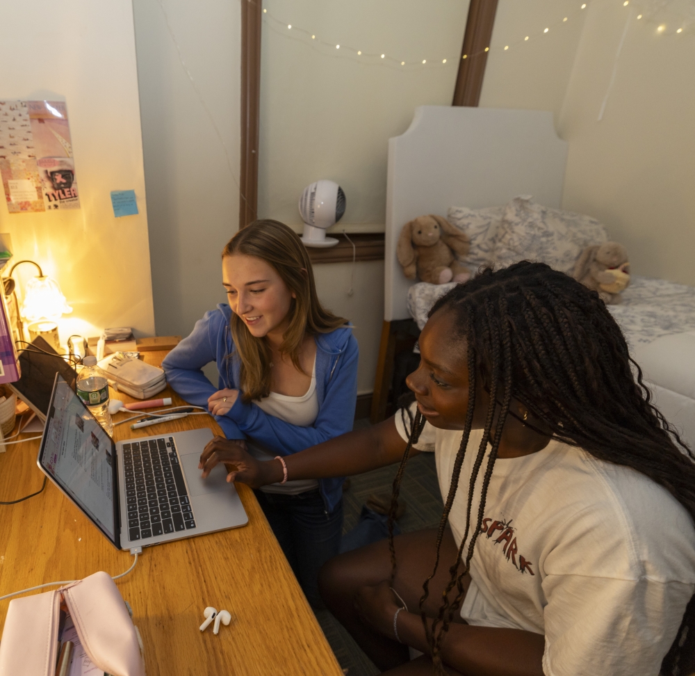Two girls look on at one's laptop in a dimly lit dorm room.