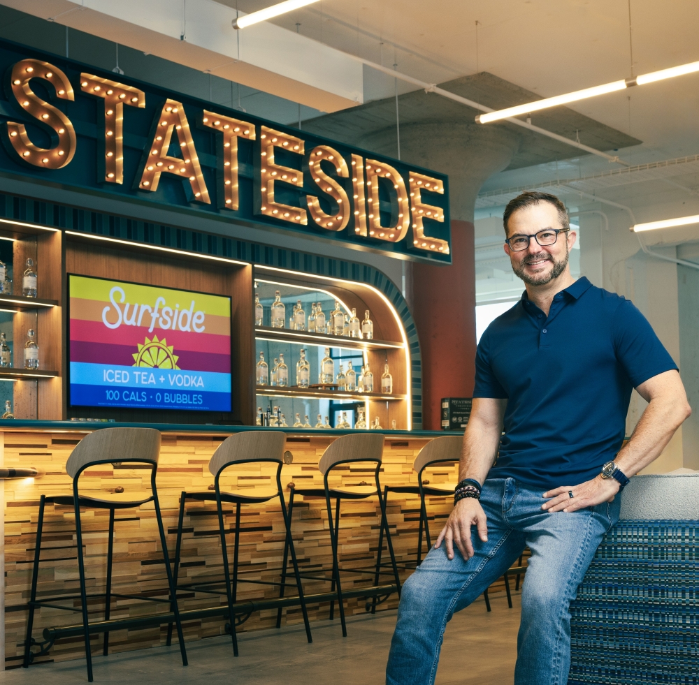 Clem Pappas, in a navy blue polo shirt and jeans, sits on the back of a blue couch with his hands resting on his upper thighs inside the bar area of his Stateside Brands headquarters. There is a long bar with a row of empty seats behind him, and in block letters formed by clear light bulbs, it spells out "STATESIDE" above the bartenders' area.