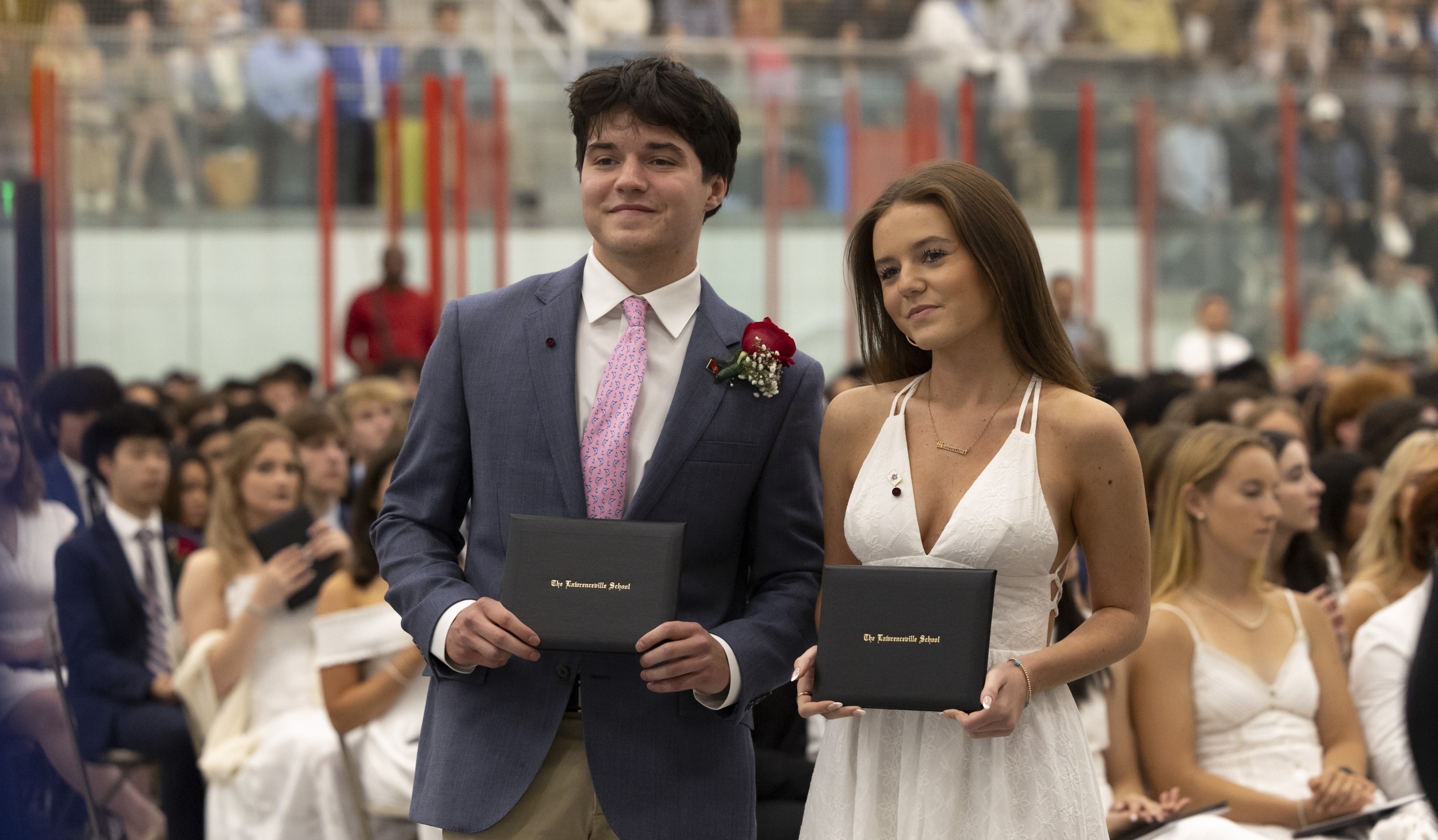 A boy in a dark blue blazer, white shirt, and pink tie, and a girl in a white dress, hold their diplomas at commencement.