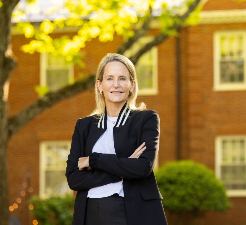 Celeste Mellet stands, smiling, with her arms folded. She is wearing a black suit and a white shirt. She has shoulder-length blonde hair and a fair complexion. Behind her is a brick academic building.