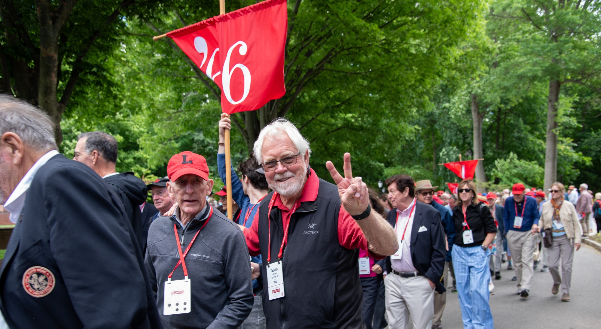 An white-haired man with a beard gives a peace sign with his left hand as he marches in the parade of alumni. There is a red flag behind him with a '66.