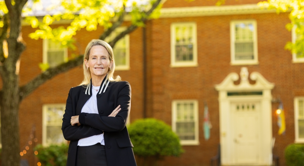 Celeste Mellet stands, smiling, with her arms folded. She is wearing a black suit and a white shirt. She has shoulder-length blonde hair and a fair complexion. Behind her is a brick academic building.