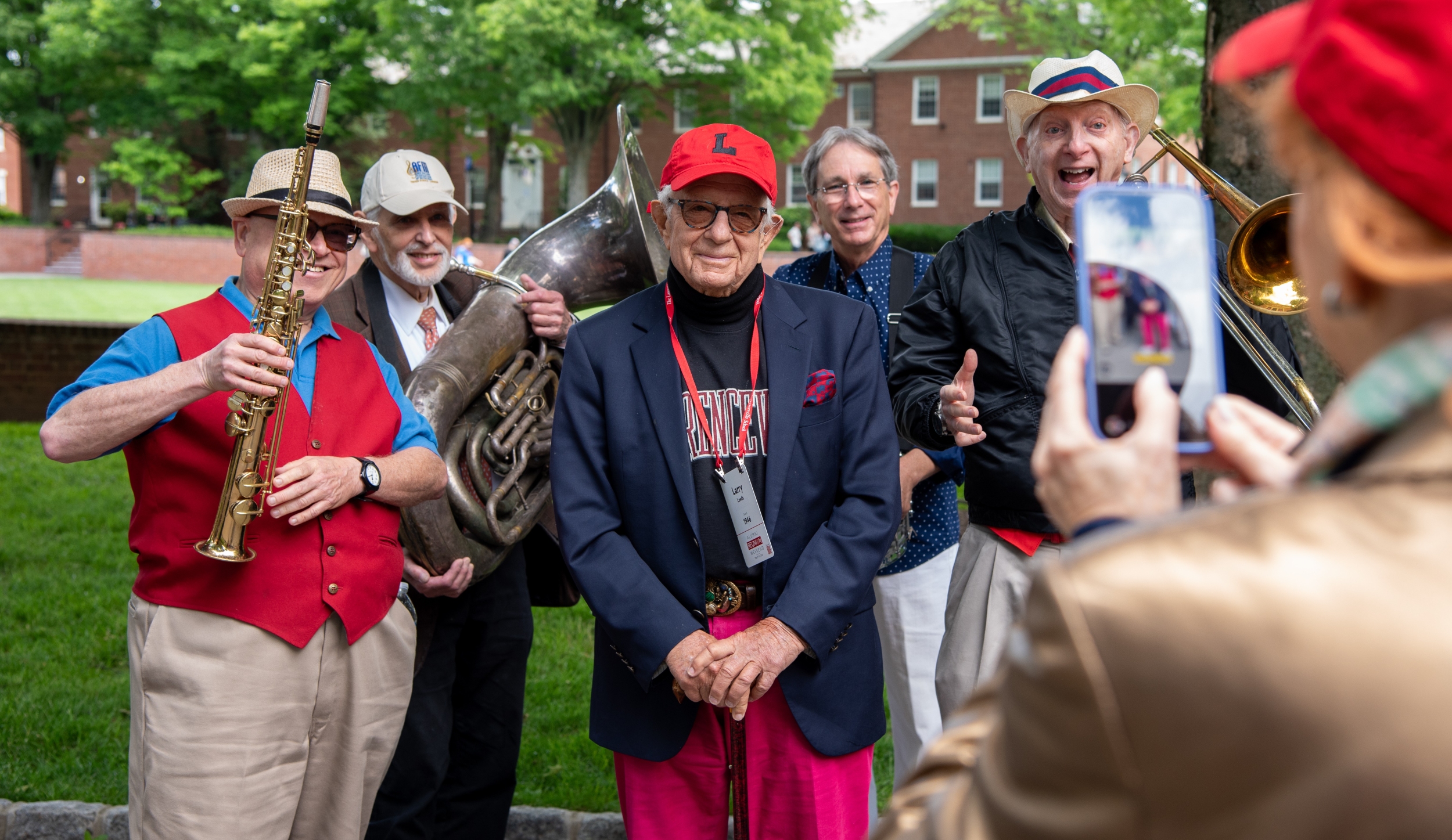 An older man wearing a red cap with a black L on it, sunglasses, red pants, and a black Lawrenceville sweatshirt beneath a navy blazer, is surrounded by members of a brass band, as they all pose for a photo.